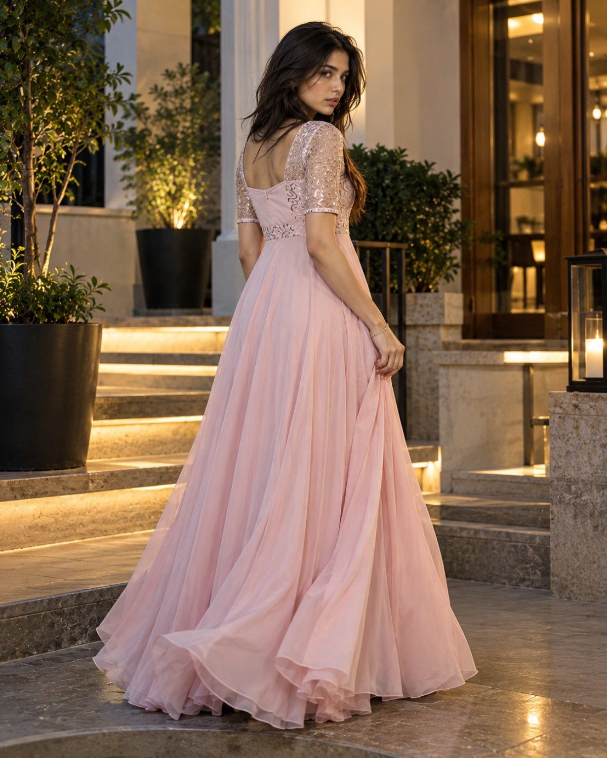 Nyra Voss in a blush-pink embellished maxi dress, photographed from behind as she turns toward the camera beside warmly lit stone steps.